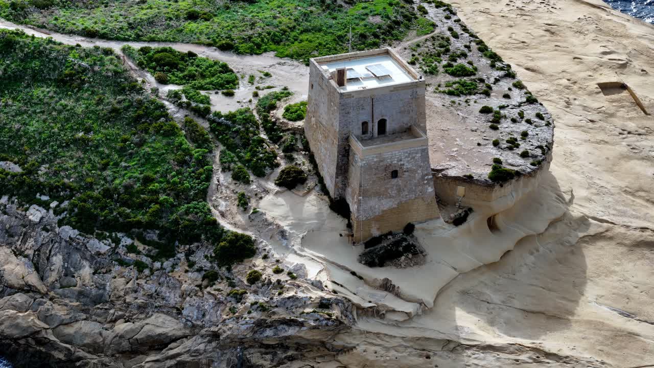 La histórica torre Xlendi se alza sobre los escarpados acantilados de piedra caliza de Gozo, Malta, durante una exuberante estación primaveral bajo un cielo despejado, con una arquitectura costera única y una vista cenital tomada con dron.