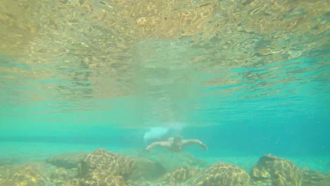 joven nadando en azul claro bajo el agua de la cascada con muchas rocas desde un ángulo plano