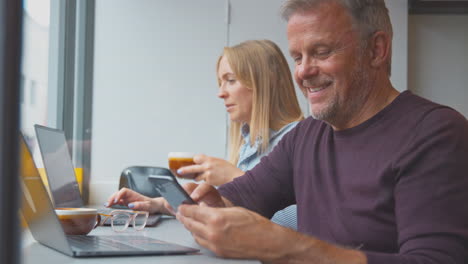 Customers-In-Coffee-Shop-Window-Working-On-Laptops-And-Browsing-On-Mobile-Phone