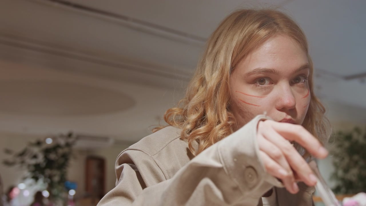 Primo piano di una giovane donna con capelli mossi che esamina e sistema la manica del suo cappotto beige in una morbida illuminazione interna. La scena presenta una calda luce ambientale e elementi dello sfondo sfocati come piante