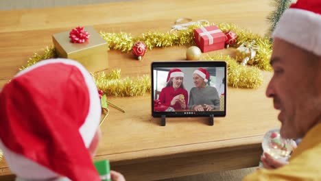 Padre-E-Hijo-Caucásicos-Con-Gorros-De-Papá-Noel-Usando-Una-Tableta-Para-Una-Videollamada-Navideña-Con-La-Familia-En-La-Pantalla