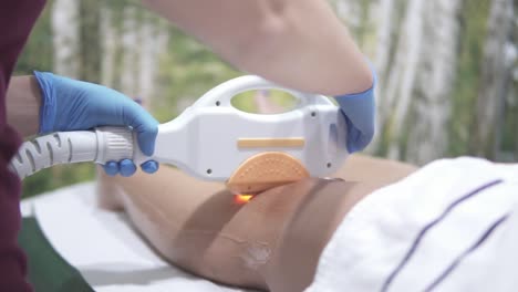 a female cosmetologist professional is using laser epilation gun to remove the hair on the anterior side of the lower leg of a young adult female caucasian client, no face closeup in slow motion