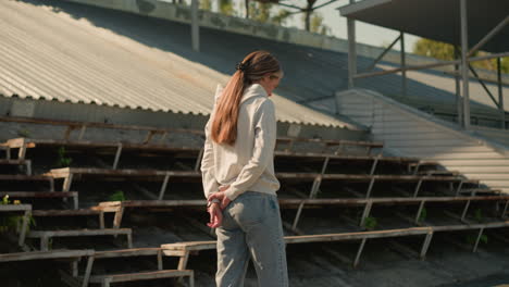 lady in hood and jeans with long hair tied back walks near empty stadium seating on a sunny day, hands behind her back, appearing deep in thought, with empty seating and surrounding greenery