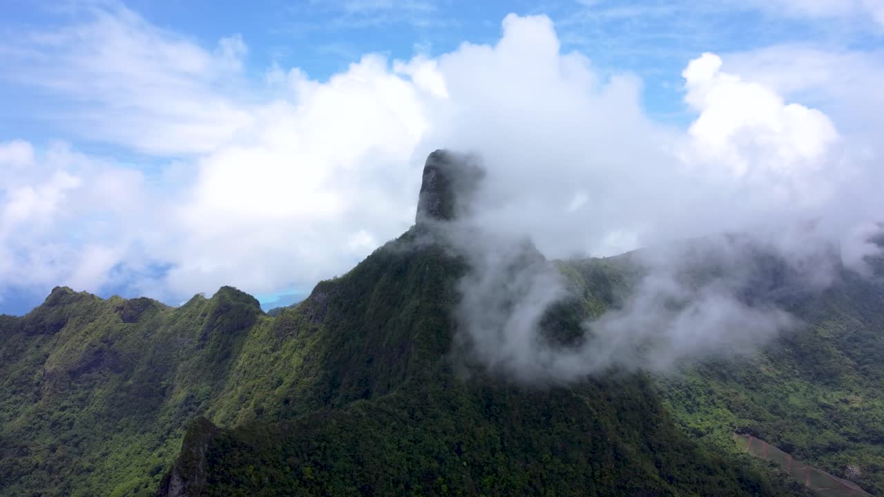 Drone view of a green tropical pacific mountain peak surrounded by clouds on a sunny day in Mouaputa, Moorea, French Polynesia.