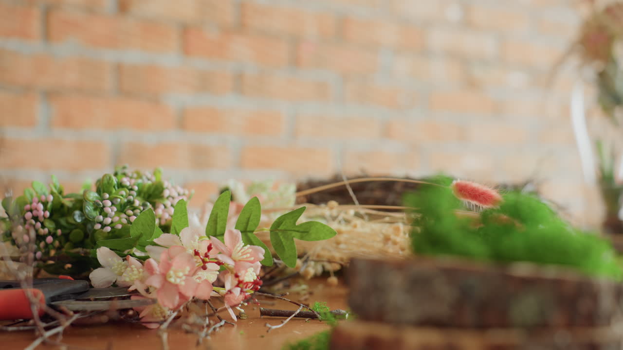 Florist hand arranging fresh greenery, pink flowers, and dried plants on wooden table with tools during creative process of making decorative floral wreath in rustic indoor workshop environment