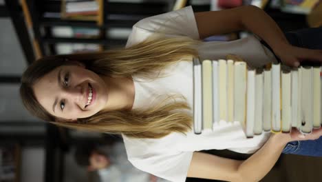 Student-girl-walks-through-library-with-stack-of-books-in-hands,-shelves-with-book,-front-view.-Young-woman-hold-books-at-hands