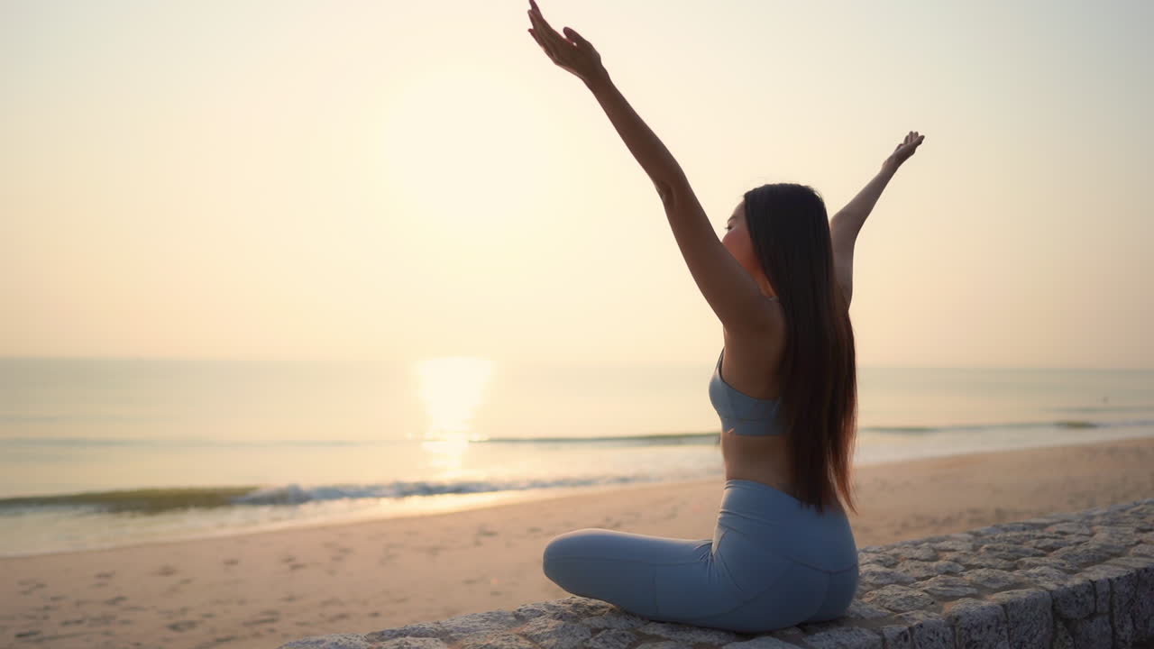 A young woman sitting on a stone wall facing the sea spreads her arms in welcome to the energy from the waves and setting sun. Copy space,