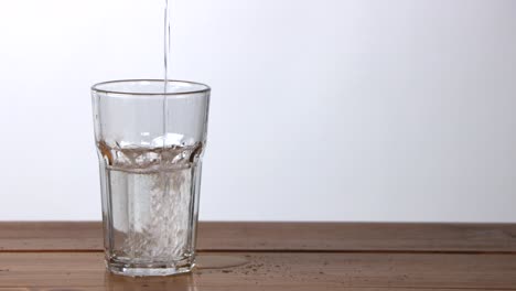 water being poured and spilled into a large glass on a wooden table with a white background