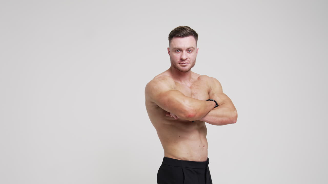 Caucasian man standing his back to camera turns around to camera. Athlete with naked torso crosses hands on his chest looking at camera. White backdrop.