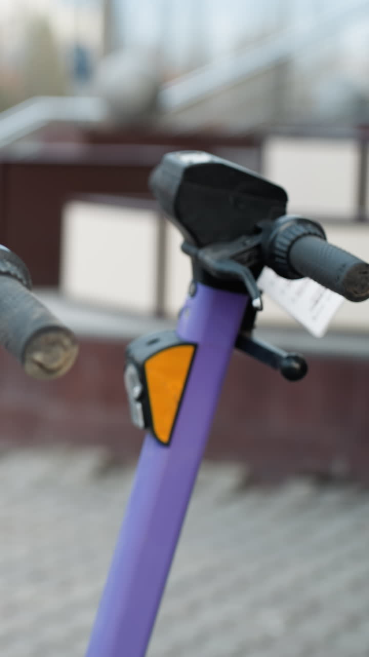 바쁜 도시 지역에 주차된 보라색 전기 스쿠터의 가까운 시각, 배경에는 움직이는 자동차와 사람들과 함께 흐릿한 도시 풍경 세부 사항이 포함되어 있습니다.