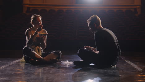 pair of weary classical ballet dancers sit on theatre stage after choreography rehearsal. ballerina eats with male dance partner after training. empty rows in theatrical hall. concept of ballet art.