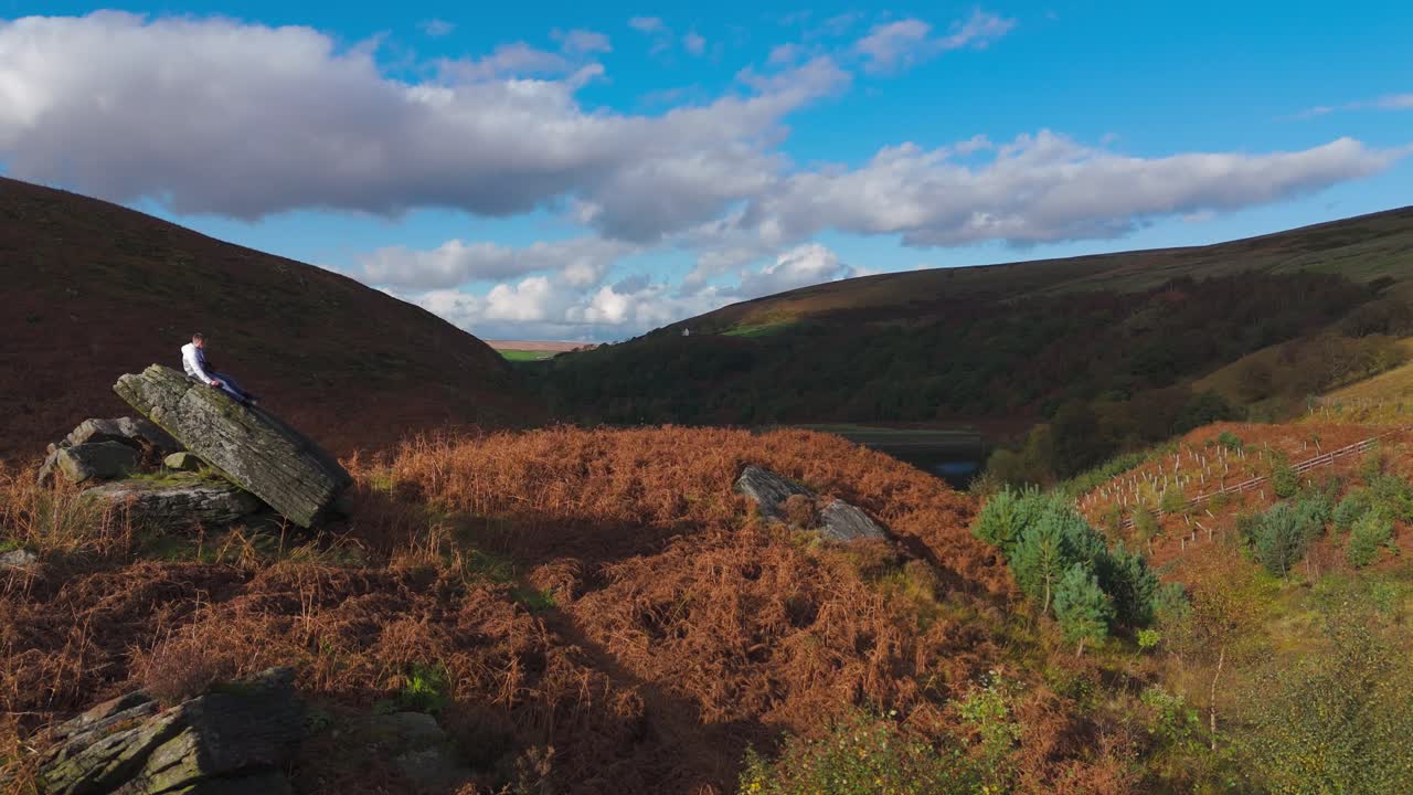 A young boy perched on a substantial rock outcrop, deep in contemplation, as aerial footage revolves around the moorland scene
