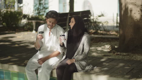 lovely muslim couple sitting on bench in park, having coffee.