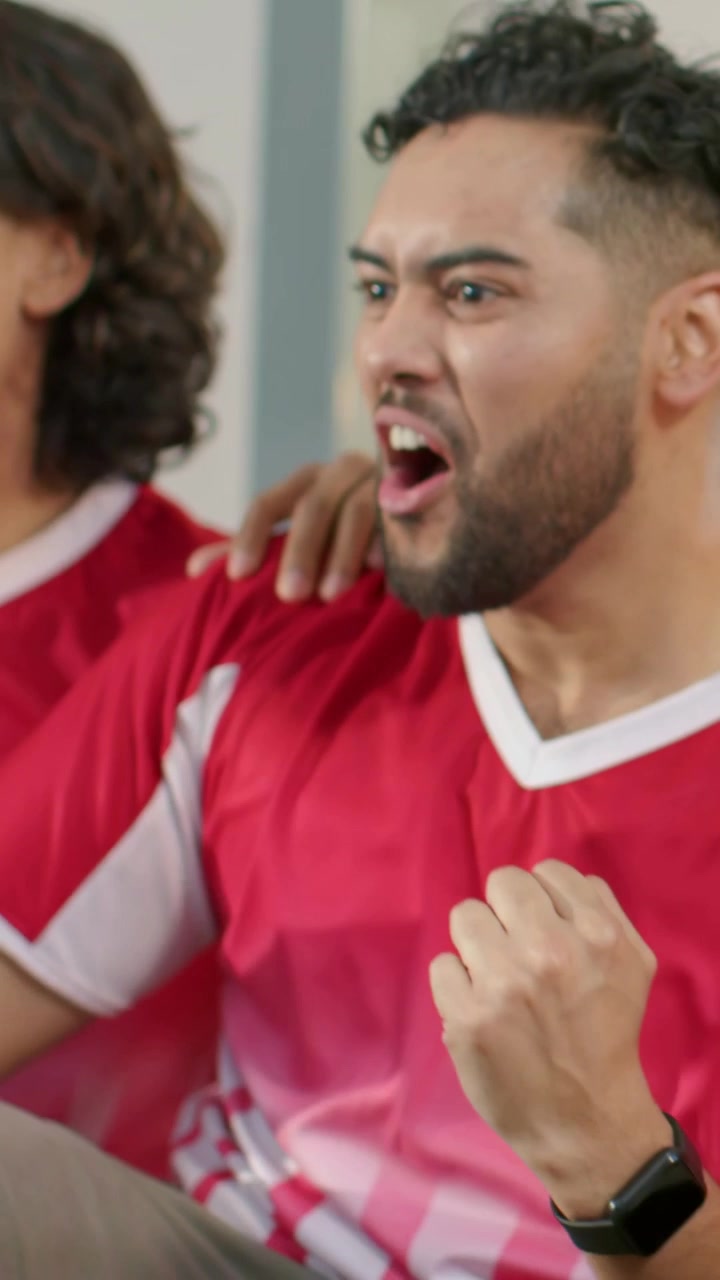 Video verticale: Reazione a un gol, donna a destra che tiene una palla, uomo che esulta sul divano con maglie rosse.