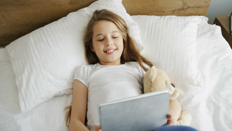 vista superior de una niña acostada en la cama por la mañana con la cara asustada mientras ve algo en la tableta