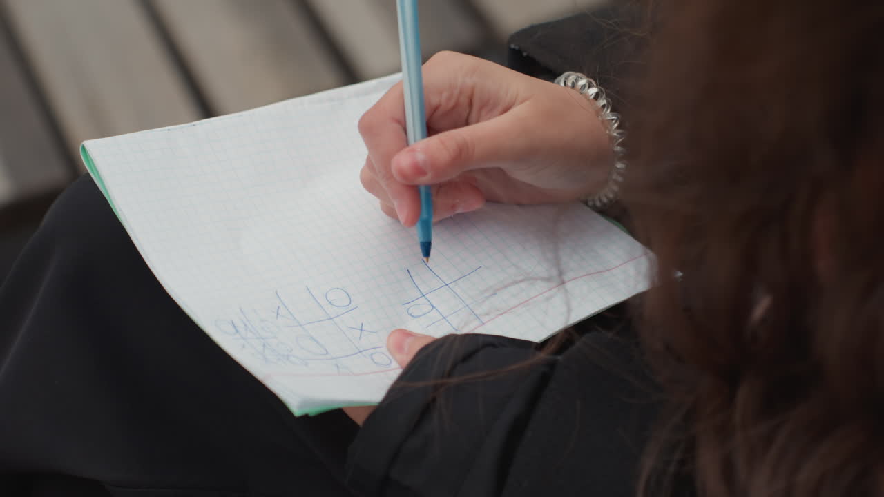 student writing notes on notebook outdoors, closeup of hand using blue pen on graph paper, bench background, bracelet visible, soft natural light, steady focused routine, candid study atmosphere