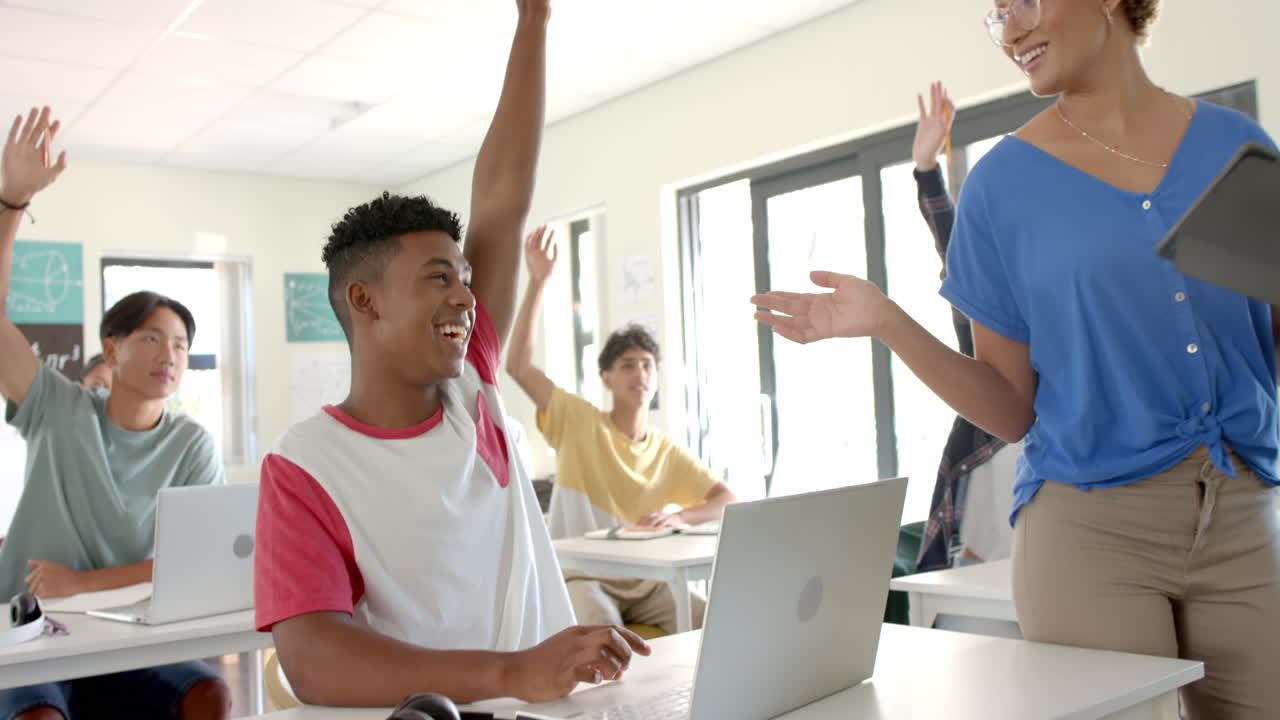 In high school, students raising hands and using laptops in classroom with teacher
