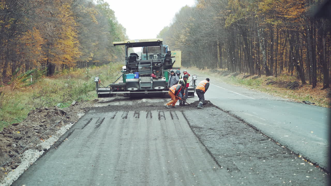 Team of workers put the hot asphalt on a half of the road on the background of a big steamroller. Road construction workers with shovels in protective uniform and the truck going by in autumn.