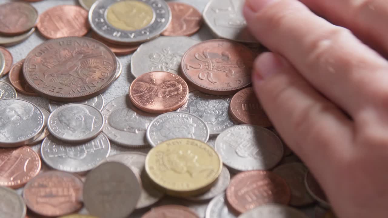 Assorted international coins from various countries and denominations, different sizes, values, colours. A man's hand spreads the pile. Slow camera movement past the coins, extreme close up