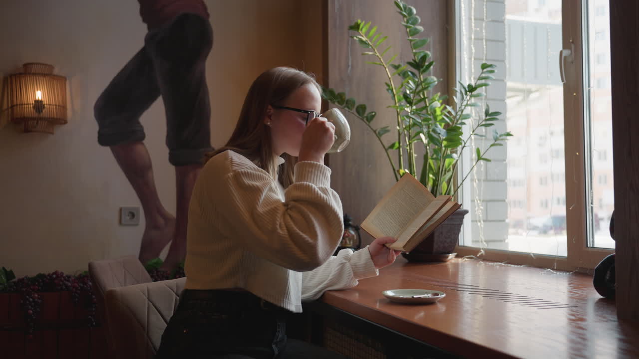 Woman in glasses reads book while sipping tea by window in cozy cafe with warm lighting, indoor plant, wall painting, and calm ambiance