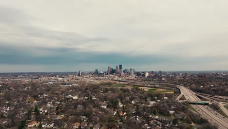 Una-Amplia-Toma-Aérea-De-Drones-Del-Horizonte-De-Minneapolis-En-Minnesota-En-Un-Día-De-Primavera-Con-Nubes-De-Tormenta-En-El-Fondo-Y-Una-Carretera-En-El-Lado-Derecho-De-La-Pantalla