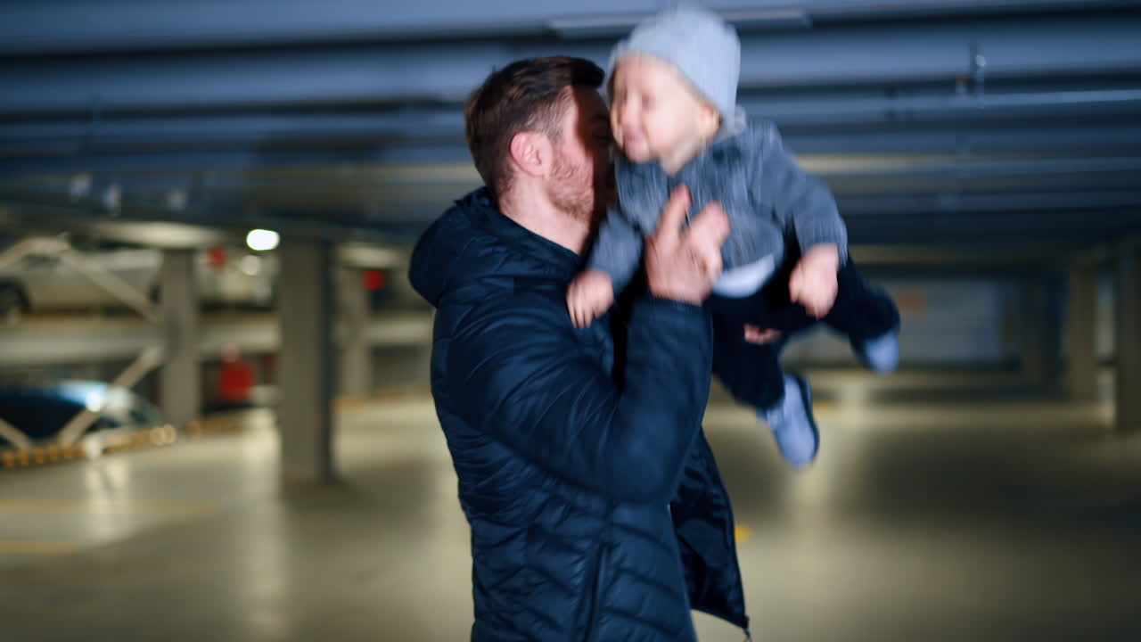 Happy Caucasian man with little infant boy in hands. Dad tosses his son and puts him on his shoulder walking away. Car park at backdrop.