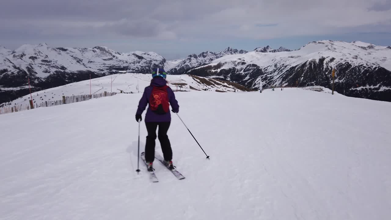 Un'inquadratura che segue una donna che scia su una pista con delle montagne sullo sfondo.