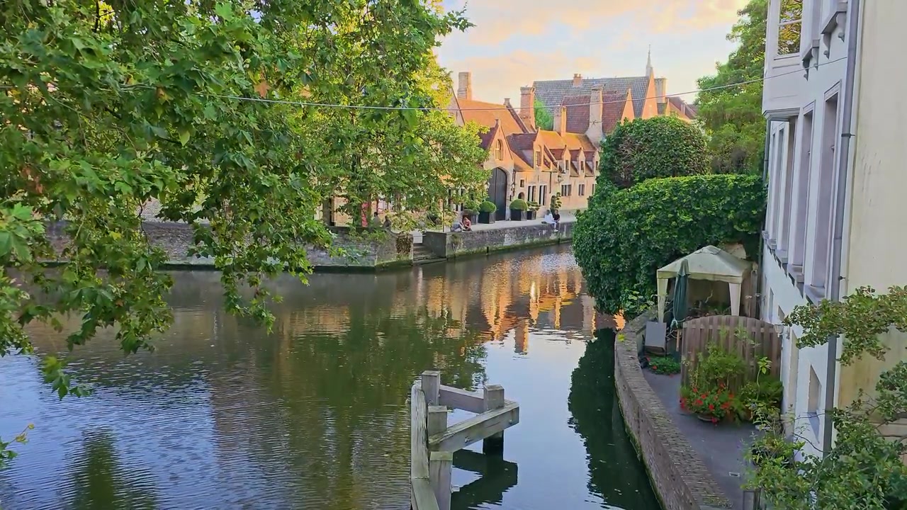 Kanalscene i Brugge, Belgien, med løvtræer, historiske murstenshuse, rolige vandrefleksioner og en stille terrasse ved floden i varmt gyldent time-lys, charmerende gammel byatmosfære.
