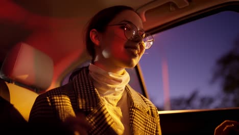 A-happy-brunette-girl-in-round-glasses-in-a-striped-white-and-black-coat-dances-after-a-happy-business-meeting-in-the-cabin-of-a-modern-car-during-a-business-trip-in-the-night-city