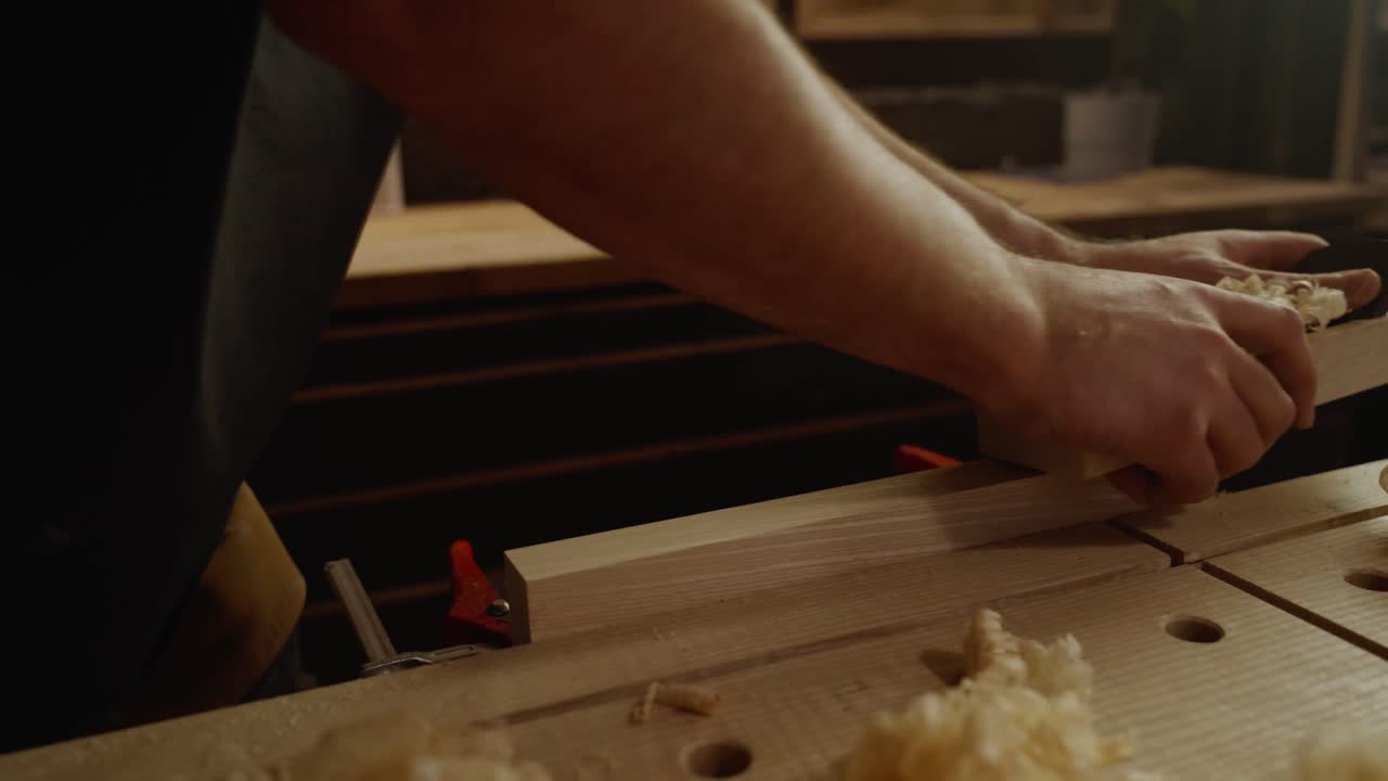 Close-up of a craftsman’s hands planing a wooden board with wood shavings scattered on the workbench, symbolizing craftsmanship, precision, and traditional woodworking