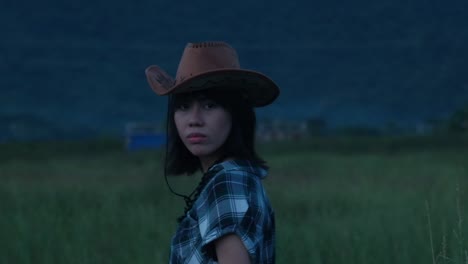rear static shot of a young asian cowgirl wearing cowboy hat dramatically turning around during moody, overcast day outdoors - slow-motion