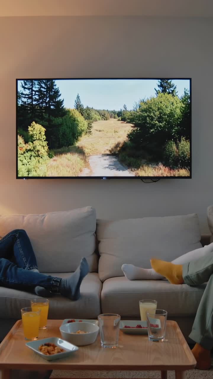 Video verticale: Coppia che si rilassa guardando la TV sul divano a casa, in jeans e abbigliamento casual verde, con succo.