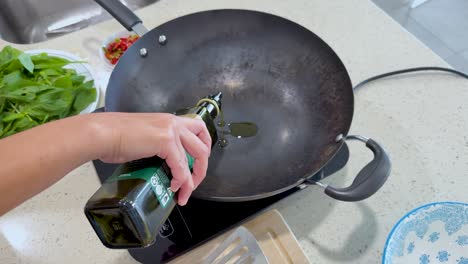a person pours olive oil from a bottle into a large black wok on an induction cooktop, surrounded by fresh vegetables in a bright kitchen