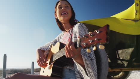 Happy-caucasian-woman-sitting-in-beach-buggy-by-the-sea-playing-guitar