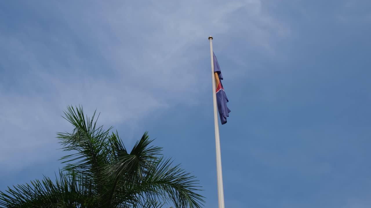 ASEAN Flag right next to a palm tree droops down on a pole while the camera slides revealing a lovely blue sky and some thin white clouds