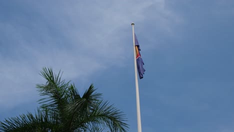 asean flag right next to a palm tree droops down on a pole while the camera slides revealing a lovely blue sky and some thin white clouds