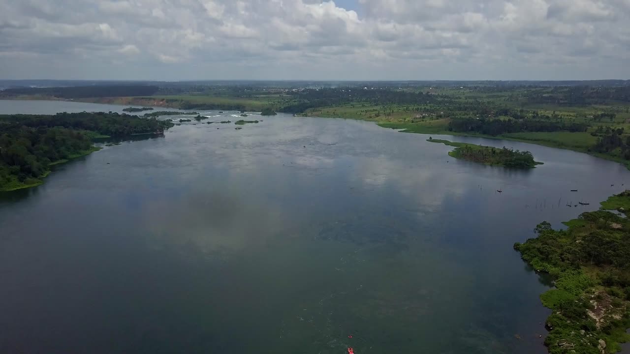 Drone pull‑out panorama of the River Nile in Uganda showing calm currents, reflective water, riverside vegetation, a small red boat and distant highland ridgelines that frame the expansive waterway