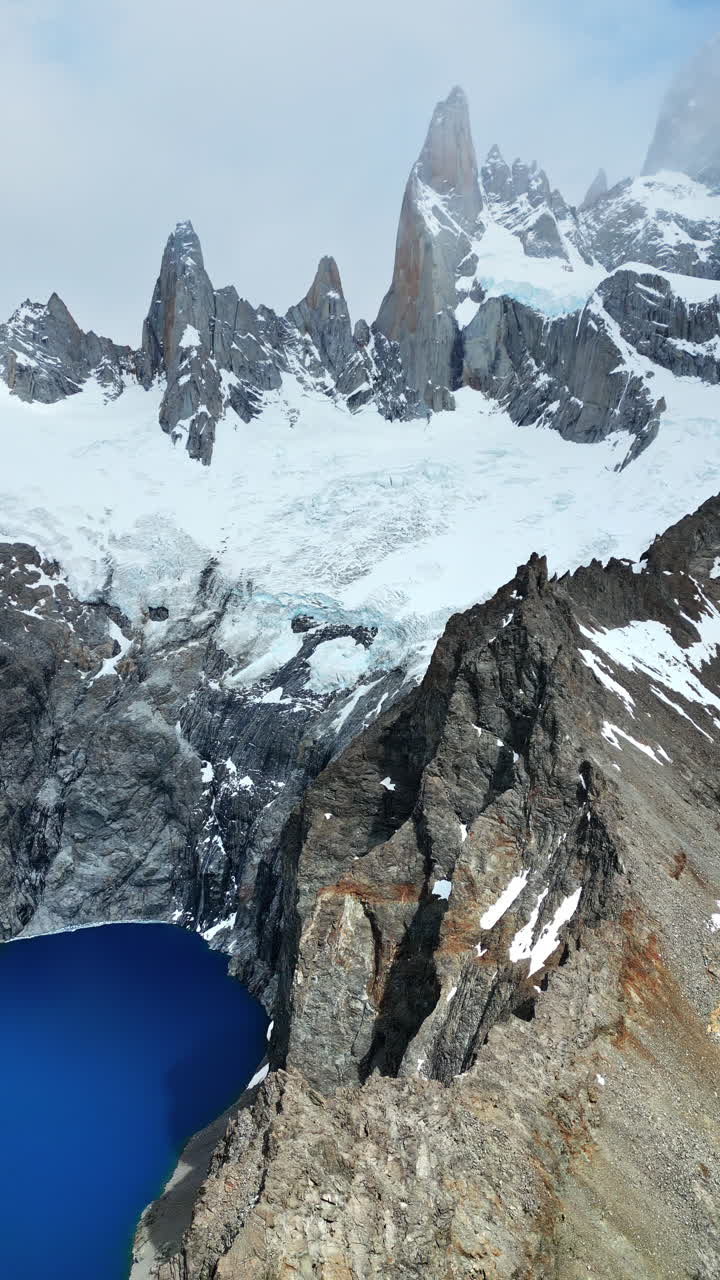 Vista aerea da drone delle imponenti cime del Fitz Roy sopra un lago glaciale blu scuro, incorniciato da scogliere rocciose e chiazze di neve. Verticale