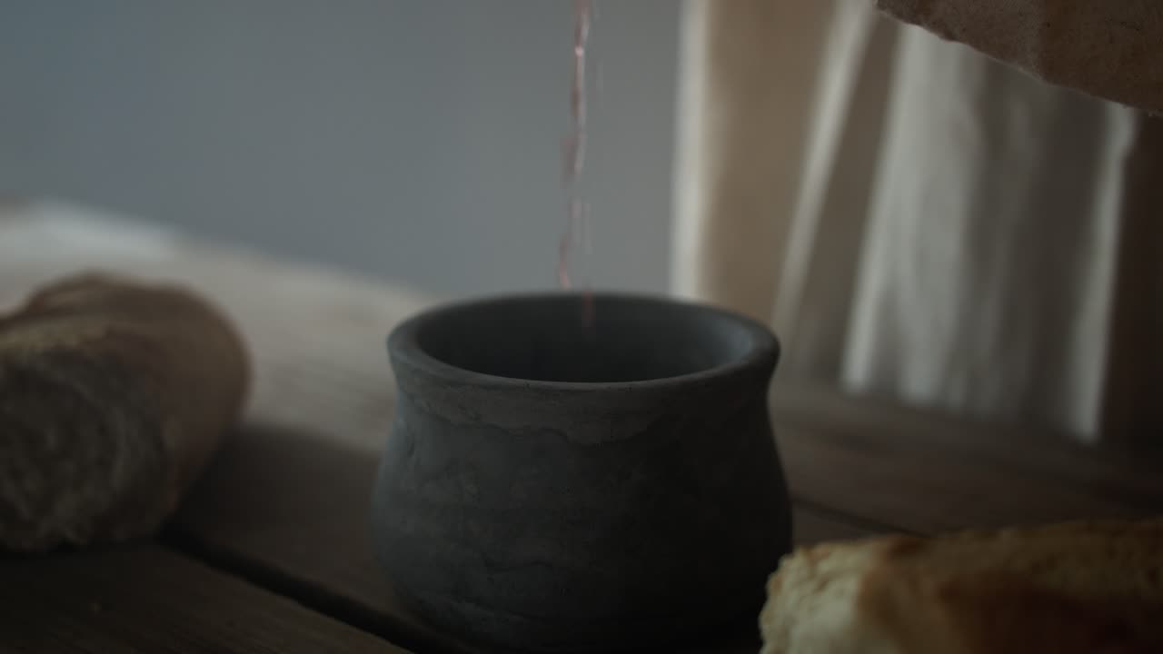 Jesus Christ pouring red wine into a clay jar on a wood table with bread in ambient, glowing light. Symbolism of the last supper, communion, Easter, Christian sacraments.