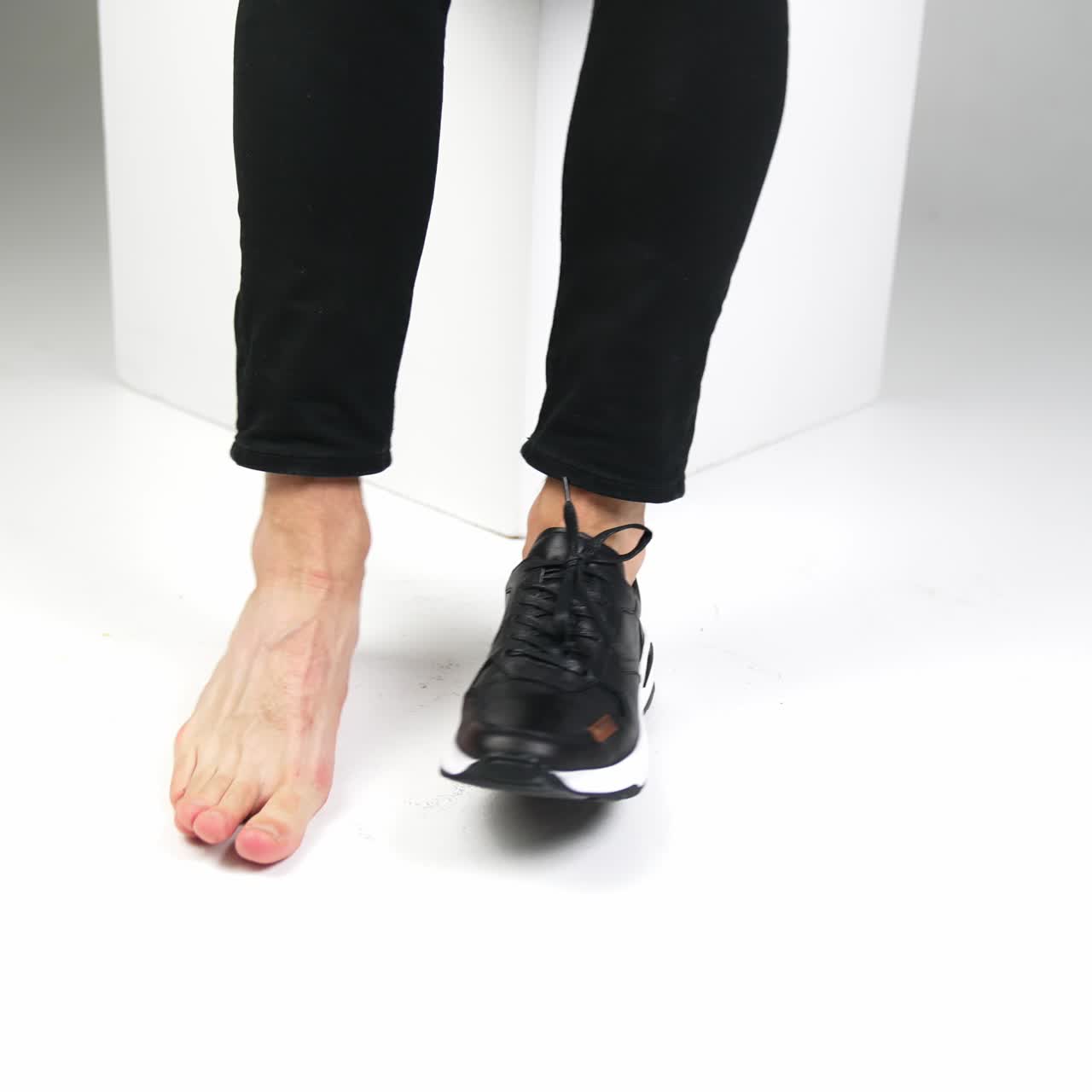 Male sitting on a white cube with one foot in a shoe and another is bare. Man moving feet on a white backdrop