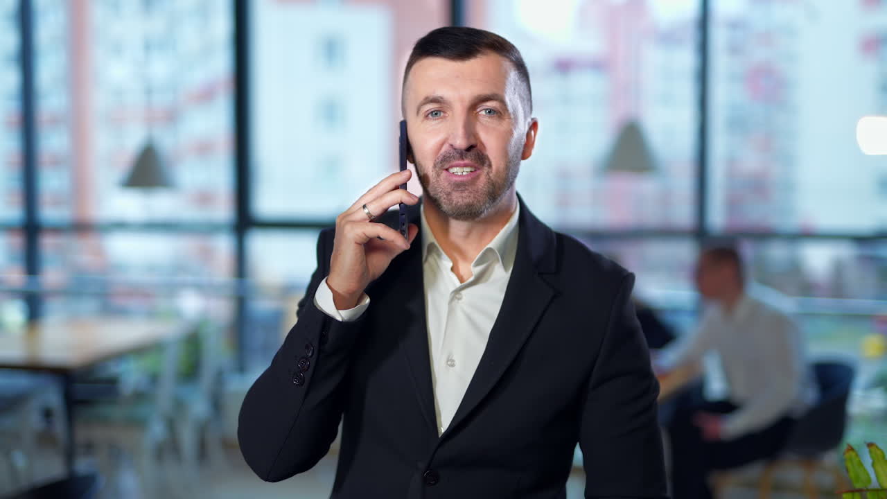 Smiling positive bearded man speaking on the phone and waving hand. Businessman looks on his watch during talk. Colleagues sit at desk at background in blur.