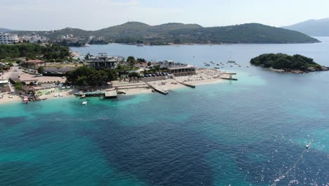 drone view in albania flying away from white sand beaches, blue crystal clear water, green small islands, boats and hotels, sunny day in ksamil