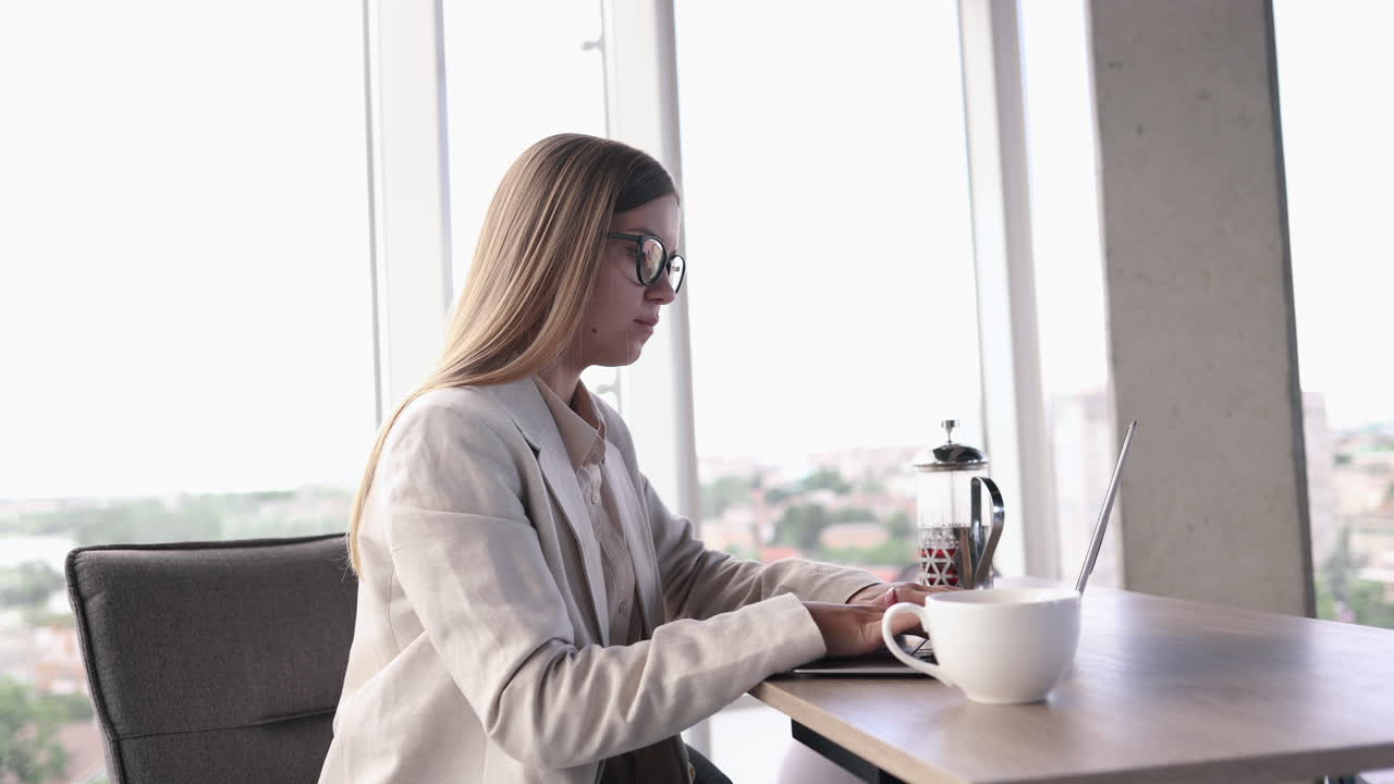 Giovane manager con gli occhiali che lavora in un ufficio luminoso e beve il tè al laptop. Un'affascinante donna dai capelli lunghi con gli occhiali alla sua postazione di lavoro.