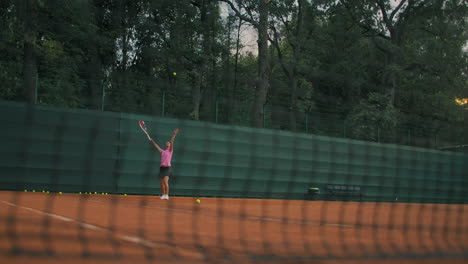 a woman in tennis training works out a smash ball and then a backhand. outdoor workout at sunset. a clay court with a green fence. wide angle through the mesh.