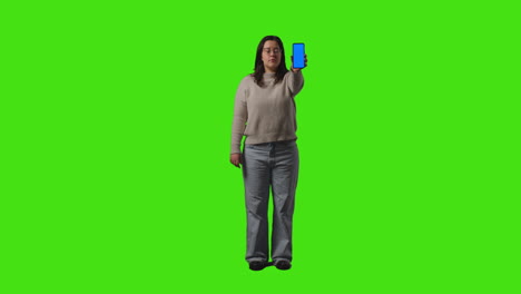 fotografía completa de una mujer con gafas sosteniendo un teléfono móvil de pantalla azul en la cámara contra un fondo de pantalla verde