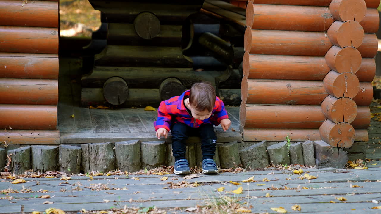 Lovely two-year-old baby boy sitting peacefully on the threshold of wooden pavilion. Cute kid stands up and runs up to camera.