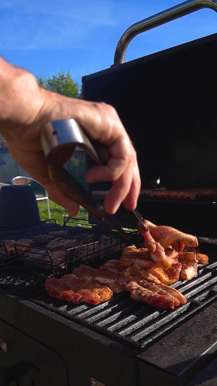 Mano di una persona che usa le pinze per girare bistecche di maiale condite che cuociono su una griglia per barbecue calda e fumante durante una giornata di sole in un giardino, con la carne che sfrigola creando un delizioso pasto all'aperto