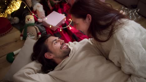 un chico moreno en un suéter blanco está tendido en el suelo en una almohada y una chica morena lo está mirando desde arriba y acostado a su lado cerca de los juguetes y regalos de año nuevo en una habitación acogedora en invierno