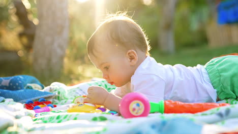 nice little caucasian toddler lies on his belly on the plaid in the garden. lovely child looking at toys in the rays of bright sun.