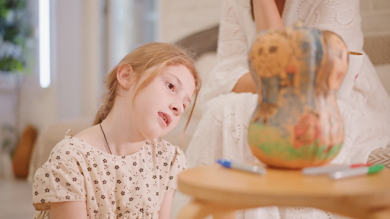 Jovem branca pintando abóbora na mesa, marcadores e esboço por perto, mãe observando do sofá, luz quente e aconchegante da sala de estar, expressão pensativa, imaginação brincalhona de torta de abóbora